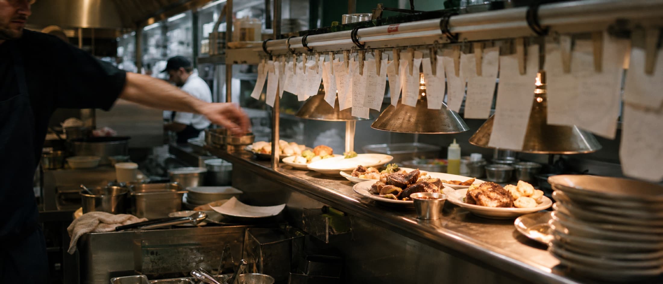 Plates waiting under heat lamps at the pass, tickets clipped to the rail above.