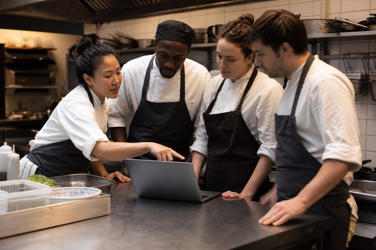 Four kitchen staff around a laptop on a prep table before service.